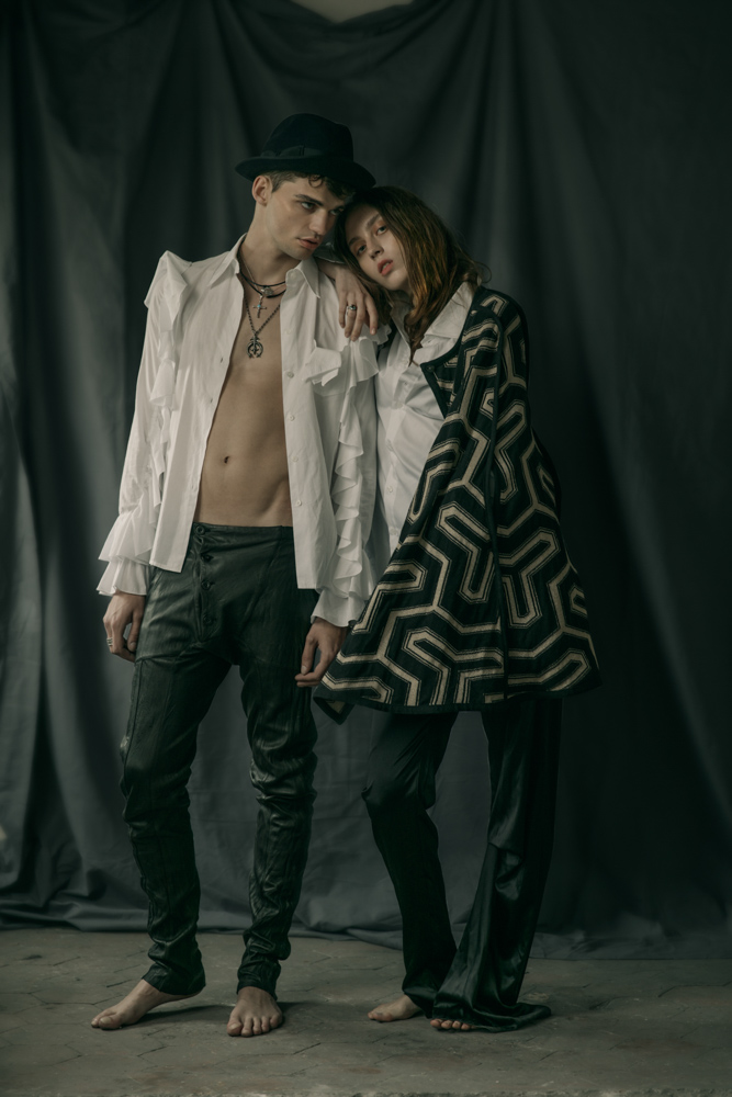 HIM White shirt with ruffles by Viktor&Rolf Leather pants by Sandrine Philippe Hat by Laurence Bossion Cross necklace, sailor wheel necklace by Harpo Gold plated metal, leather necklace from Nature collection by Delphine-Charlotte Parmentier Ring by Harpo HER White shirt by Hugo Boss Asymmetrical black trousers by Patuna Print knit cape by Maria Aristidou Necklace, ring by Harpo